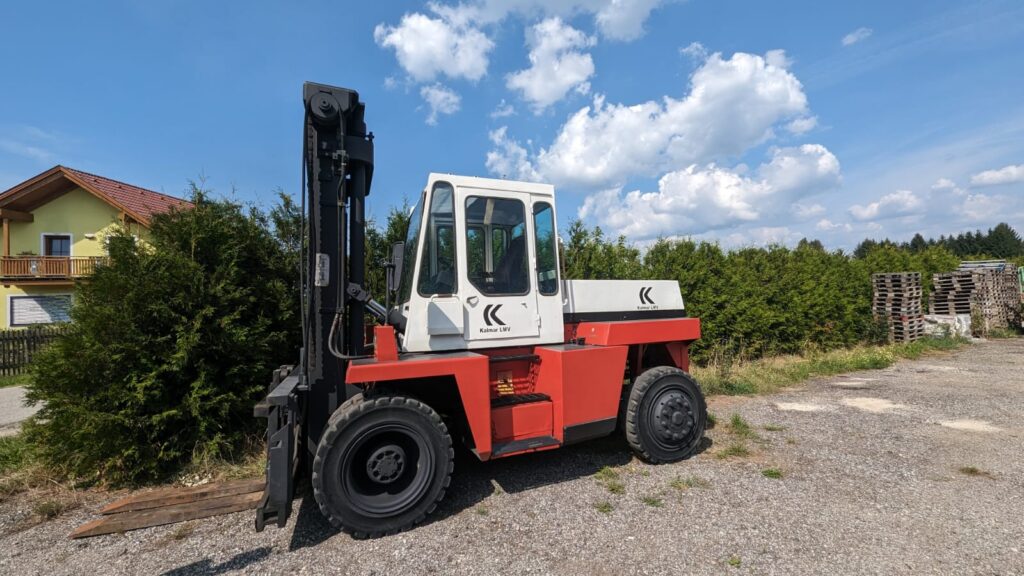 Ein roter und weißer Gabelstapler steht auf einem Schotterplatz mit Bäumen im Hintergrund.